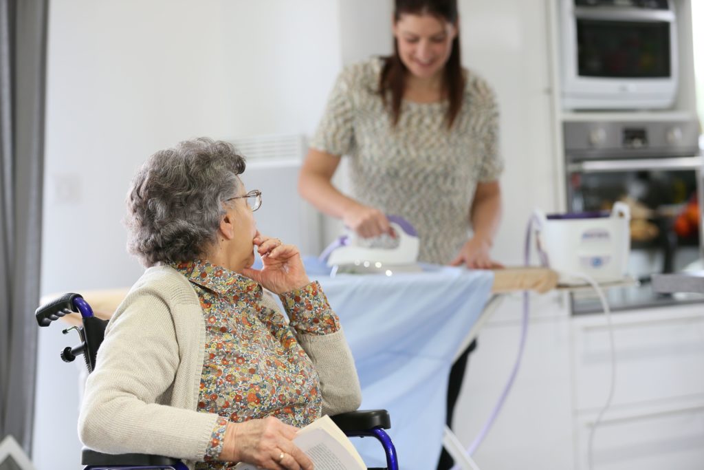 femme âgée assise dans un fauteuil roulant regardant son aide à domicile qui repasse une chemise