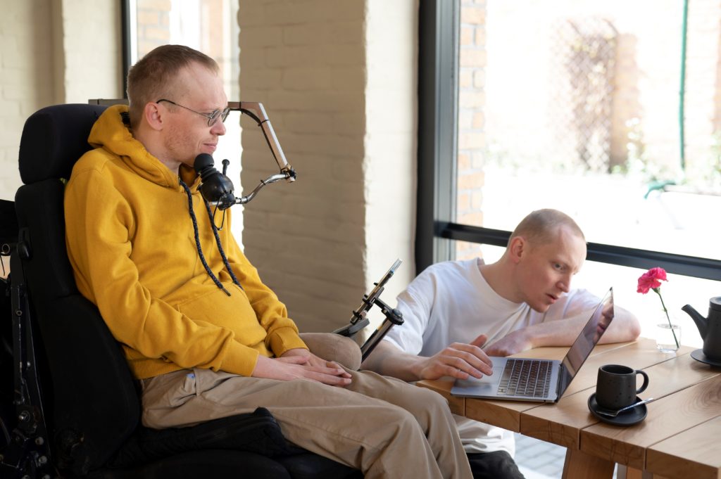 auxiliaire de vie aidant un homme en situation de handicap assis dans un fauteuil roulant électrique à utiliser son ordinateur