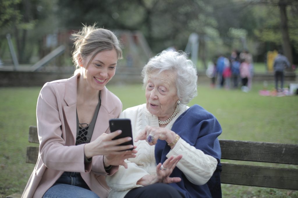 aide à domicile aidant une femme agée à utiliser son smartphone, assises sur un banc dans un parc